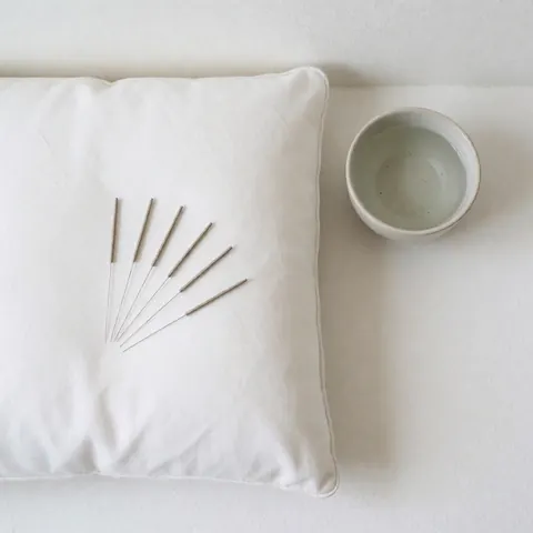 Aiguilles d'acupuncture disposées proprement sur un fond blanc minimaliste, accompagnées d'un coussin et d'une tasse d'eau pour le bien-être.