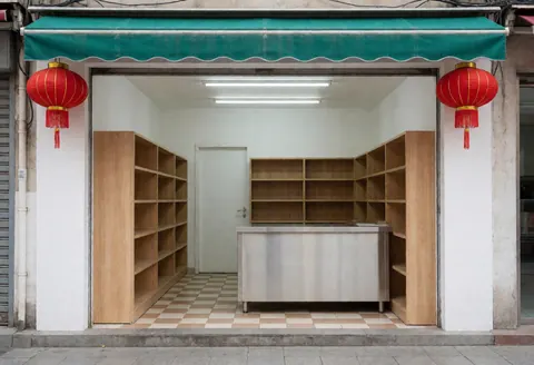 Entrée de minimarché avec murs blancs, sol carrelé, étagères en bois et comptoir en acier inoxydable, deux lanternes rouges suspendues et auvent vert, ambiance propre et invitante.