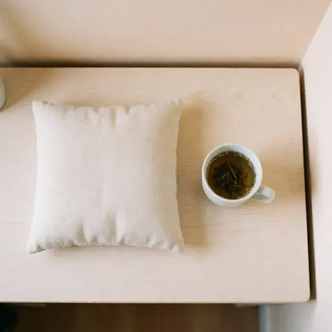 Composition zen avec un coussin doux, une tasse de thé fumant posée sur une surface en bois clair, évoquant la détente et le bien-être.