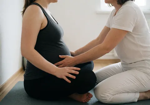 Une femme enceinte reçoit un massage doux du bassin d'une professionnelle. Scène de préparation à l'accouchement dans une pièce lumineuse aux murs blancs.