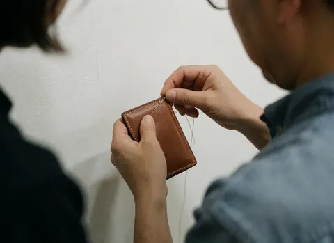Gros plan sur les mains d'un artisan coudant un portefeuille en cuir avec une aiguille et du fil, mettant en valeur la texture du matériau et la précision de la couture.