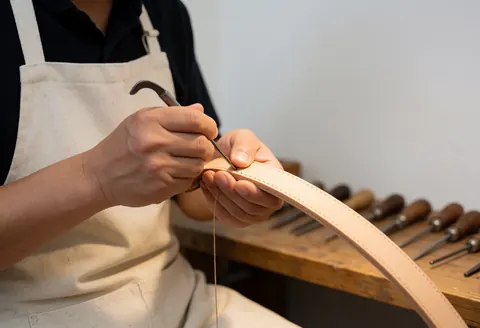 Mains d'un artisan coudant du cuir avec une aiguille et un poinçon sur un établi, outils visibles, éclairage chaud et fond blanc.
