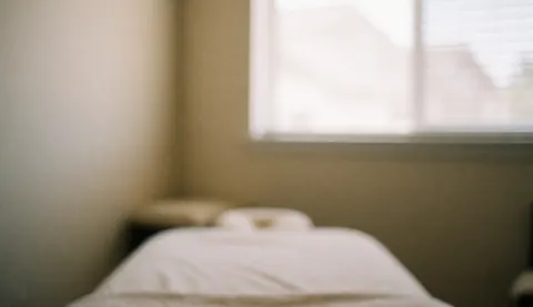 Vue d'une pièce calme avec un lit de massage blanc, des murs beige clair et une fenêtre équipée de stores, servant de fond neutre pour le design.