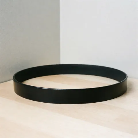 Ceinture en cuir noir aux bords lisses et brillants posée sur une surface en bois clair, avec un arrière-plan gris et une éclairage professionnel.