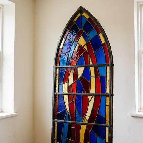 Fenêtre en vitrail gothique avec des verres bleus, rouges et dorés, installée dans un atelier aux murs blancs et à la lumière naturelle.