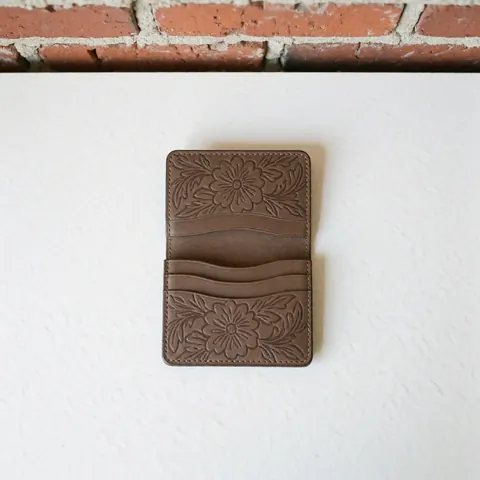 Vue de dessus d'un portefeuille en cuir brun orné de motifs floraux brodés, posé sur un fond blanc et des briques rouges. Produit fini au design artisanal avec texture visible.