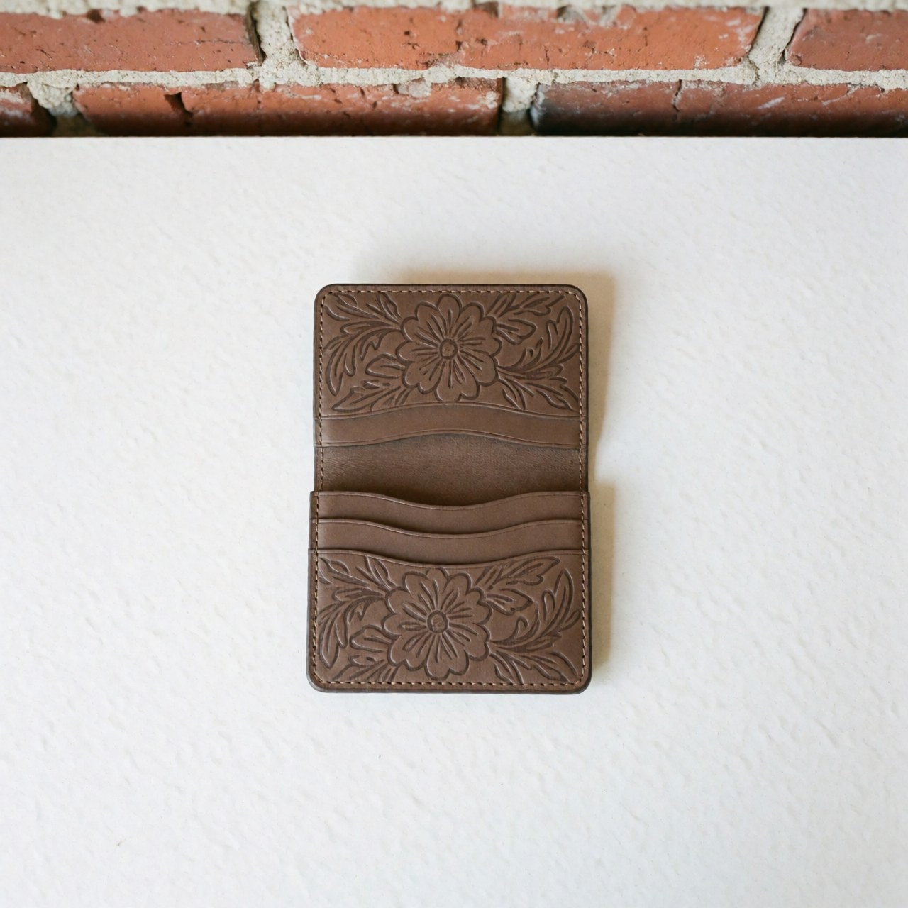 Vue de dessus d'un portefeuille en cuir brun orné de motifs floraux brodés, posé sur un fond blanc et des briques rouges. Produit fini au design artisanal avec texture visible.