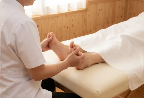 Un thérapeute en uniforme blanc effectue un massage des pieds sur un client allongé sur une table avec drap blanc dans un espace lumineux aux murs en bois.