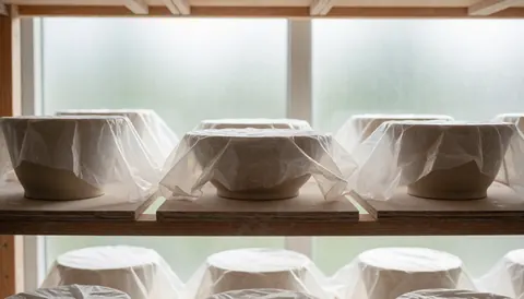 Des pièces de céramique en cours de séchage sont protégées par un film plastique transparent, posées sur des étagères en bois dans un environnement lumineux et calme.