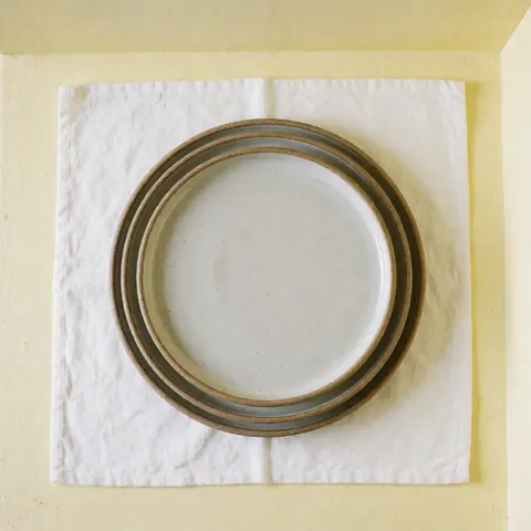 Des assiettes en céramique artisanale sont empilées sur un tissu blanc, avec des murs de couleur jaune clair, une illumination naturelle et propre.