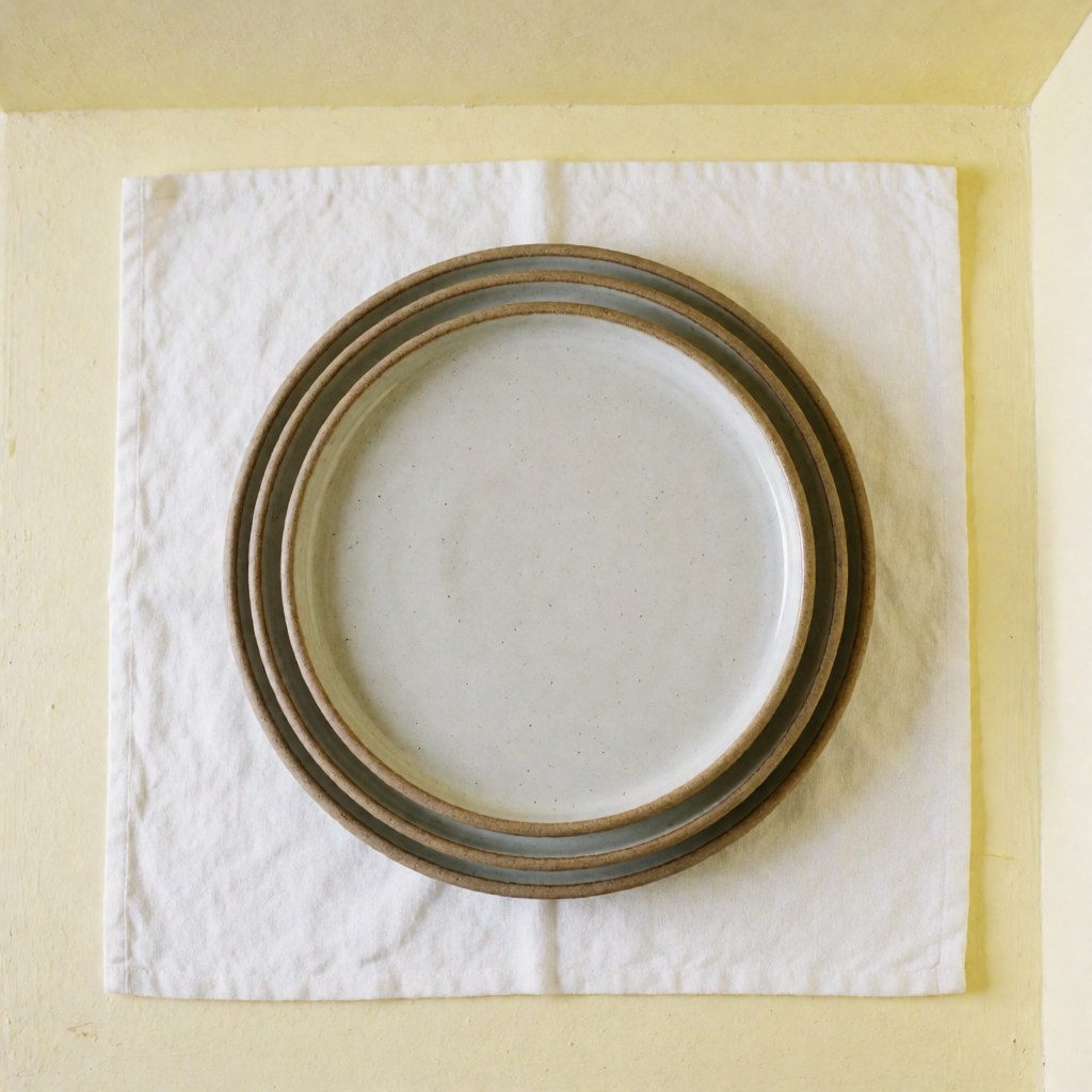 Des assiettes en céramique artisanale sont empilées sur un tissu blanc, avec des murs de couleur jaune clair, une illumination naturelle et propre.