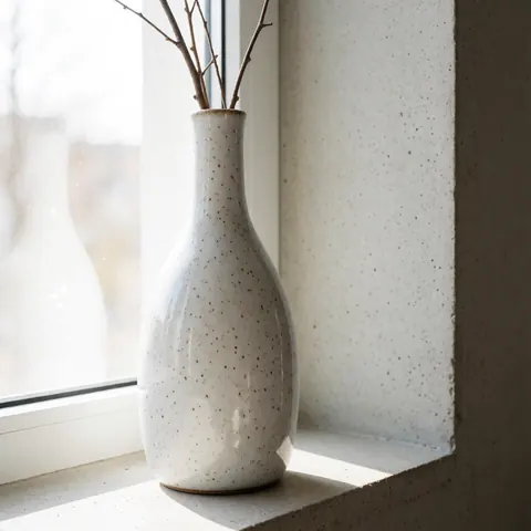 Un vase en céramique artisanale à la surface mouchetée et glacée, posé sur un mur de béton, orné de petites brindilles sèches, baigné de lumière naturelle.