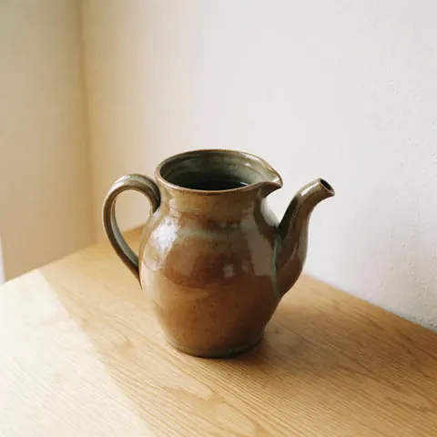 Un vase d'eau en céramique artisanale de couleur marron avec un émail brillant, posé sur une surface en bois clair devant un mur blanc.