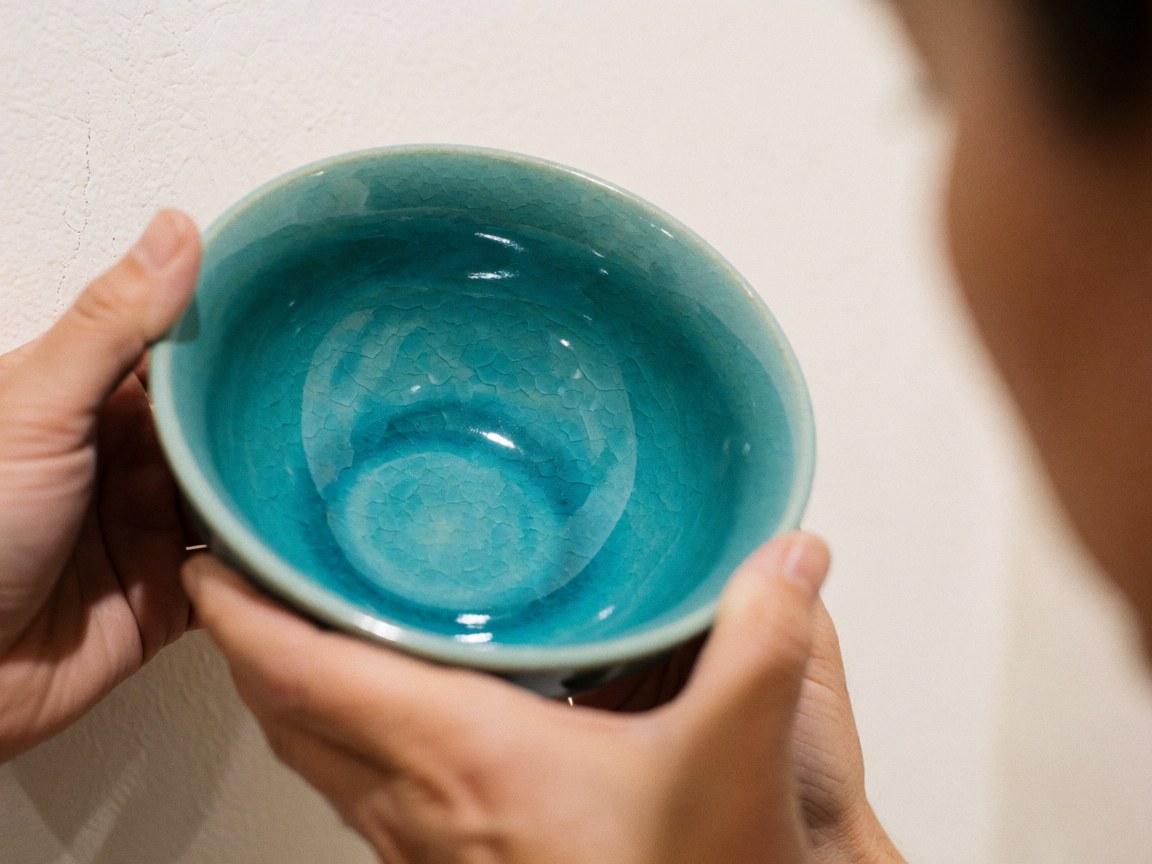 Bol artisanal en céramique au glaçage turquoise vif, tenu par des mains, sur un fond de mur chaud. Idéal pour la décoration et l'utilisation en cuisine.