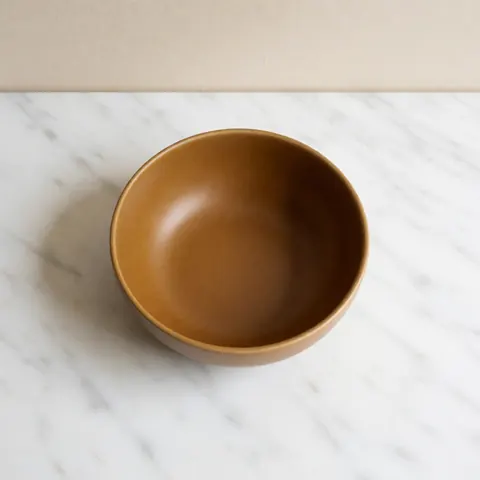 Bol en céramique artisanale aux tons ocres posé sur une surface en marbre blanc avec un fond beige et un éclairage doux, idéal pour une table à manger moderne.