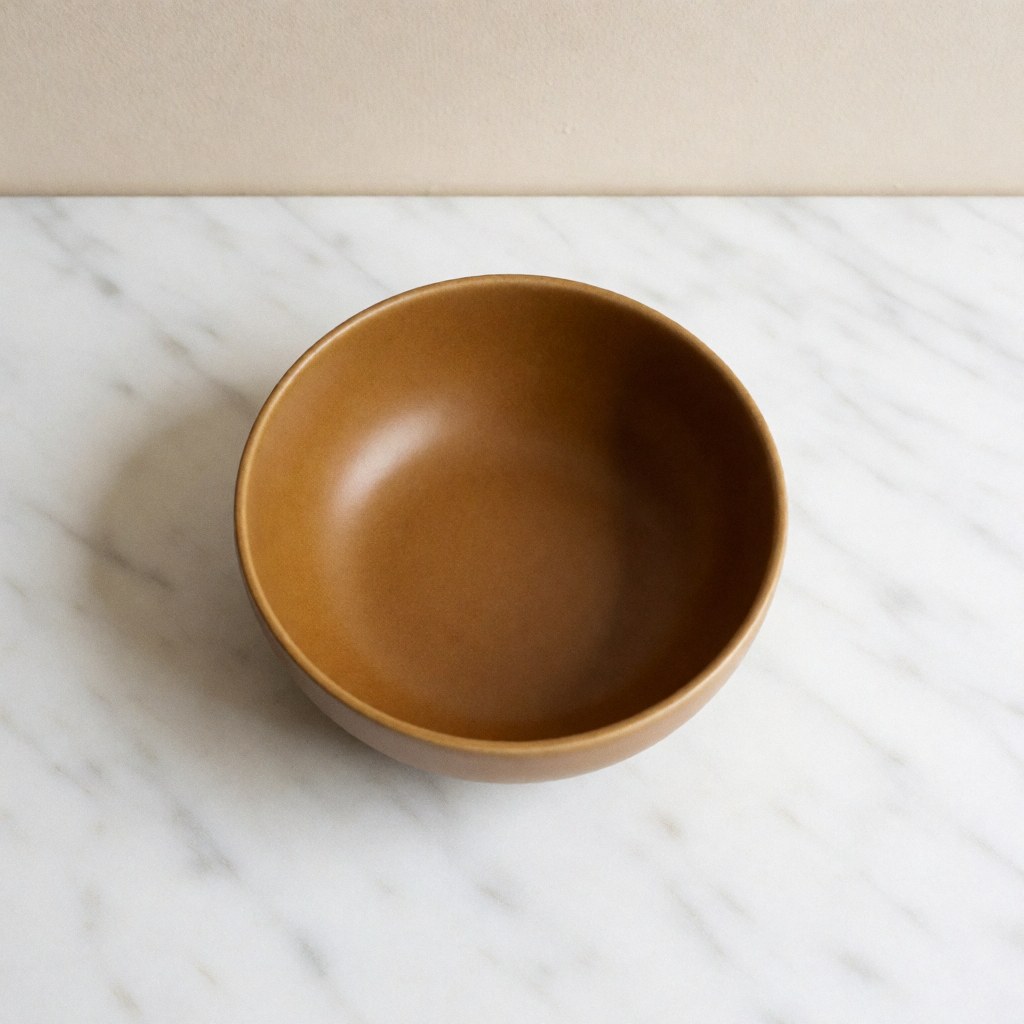 Bol en céramique artisanale aux tons ocres posé sur une surface en marbre blanc avec un fond beige et un éclairage doux, idéal pour une table à manger moderne.