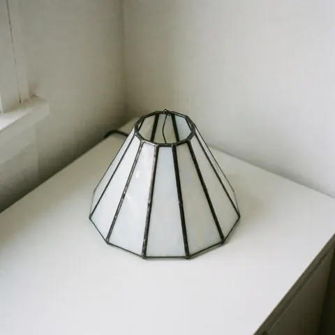 Lampe de style minimaliste avec un corps en verre blanc translucide et un cadre métallique fin, posée sur une surface claire près d'un mur blanc.