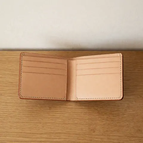 Un portefeuille en cuir marron déplié révélant son intérieur avec des compartiments et des fentes pour cartes, posé sur une surface en bois clair.