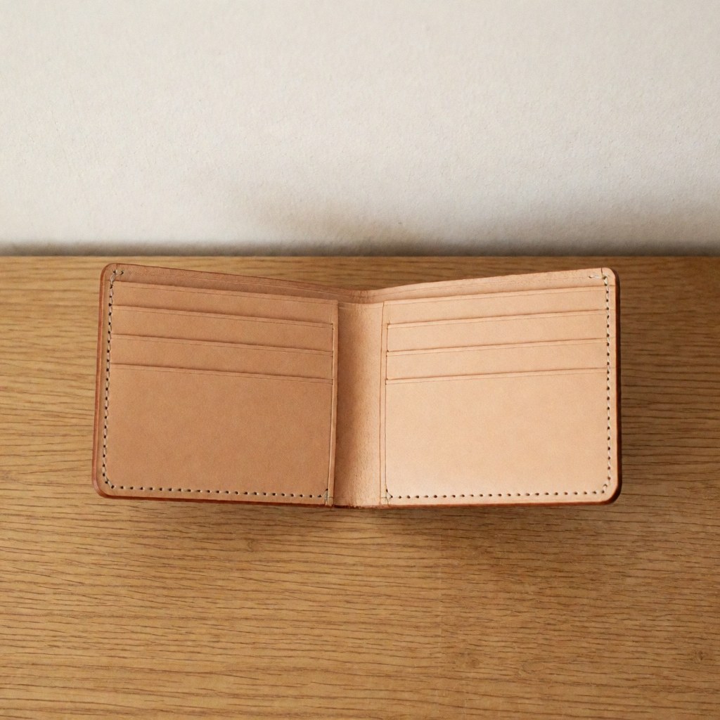 Un portefeuille en cuir marron déplié révélant son intérieur avec des compartiments et des fentes pour cartes, posé sur une surface en bois clair.