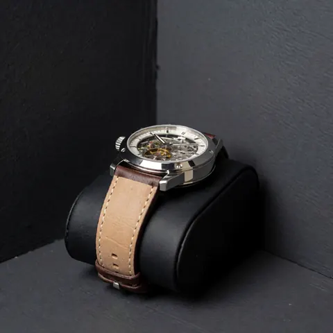 Montre-bracelet mécanique à bracelet en cuir, mouvement visible à travers le verre, sur coussin noir, murs gris, éclairage doux.