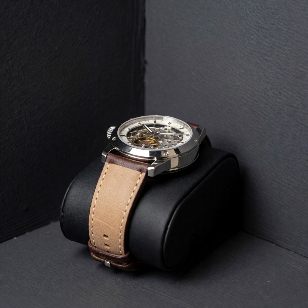 Montre-bracelet mécanique à bracelet en cuir, mouvement visible à travers le verre, sur coussin noir, murs gris, éclairage doux.