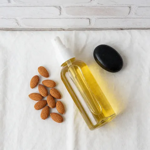 Bouteille d'huile d'amande avec spray, amandes entières et pierre noire sur tissu blanc, idéale pour massages et spa.