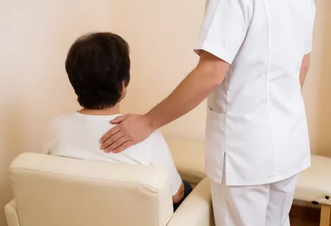 Un professionnel en uniforme blanc effectue une évaluation sur l'épaule d'un client assis dans un cadre de soin calme.
