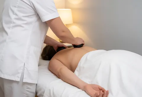 Un professionnel en uniforme blanc applique des pierres chaudes sur le dos d'un client allongé sur une table recouverte de serviettes blanches dans un cadre apaisant.