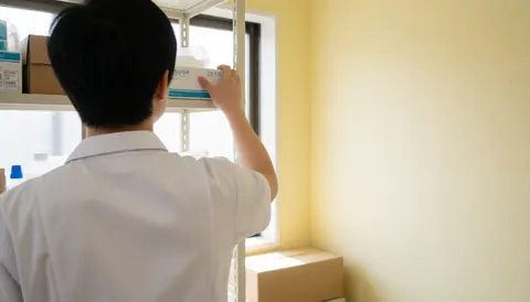 Dos d'un professionnel en uniforme rangeant des boîtes sur une étagère dans un cabinet médical, avec lumière naturelle et mur jaune.