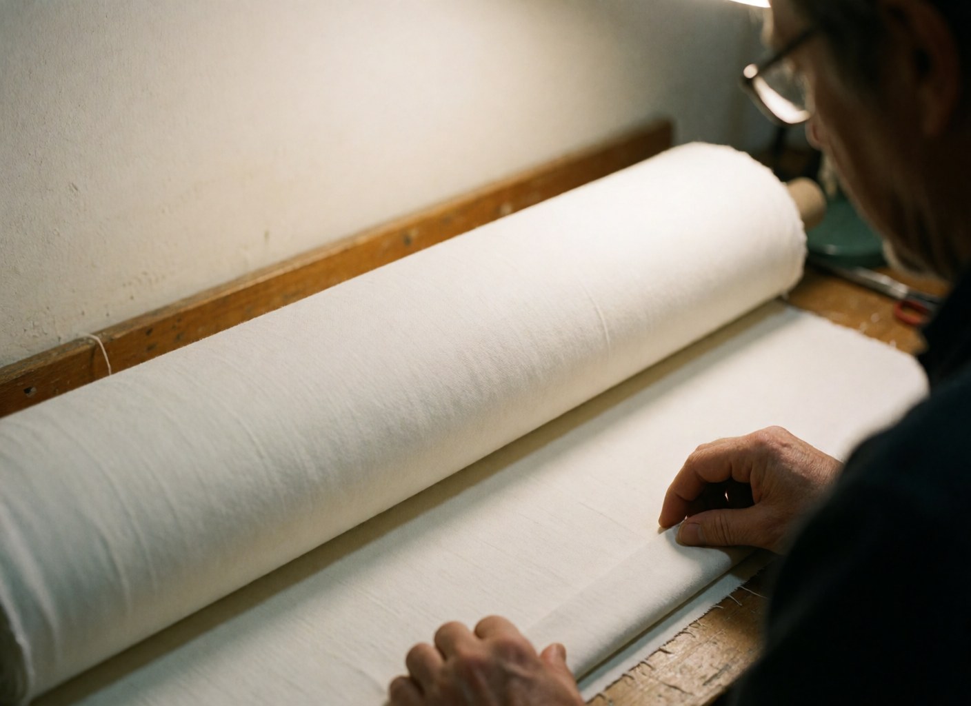 Rouleau de tissu blanc sur un établi en bois avec des mains travaillant, éclairage chaleureux et murs blancs pour textile artisanal.
