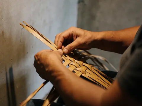 Mains d'un artisan tissant avec précision des fibres de palmier sec sur un fond de mur en béton, illustrant une scène d'artisanat traditionnel.
