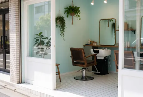 Fauteuil de barbier, vasque blanche, miroir en bronze et plantes suspendues dans un salon aux murs turquoise et sol blanc, vu depuis la rue.