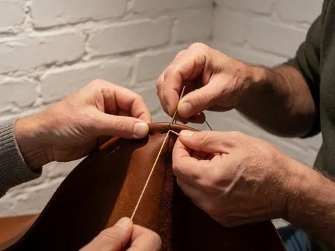 Gros plan montrant des mains travaillant le cuir avec une aiguille et du fil, mettant en évidence la texture du matériau et les outils de travail sur un fond neutre.
