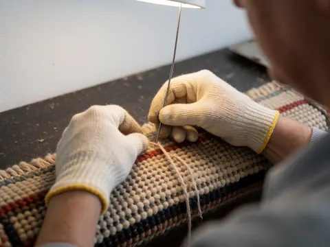 Gros plan d'une main portant un gant de travail en train de coudre ou réparer un tapis à la main, mettant en valeur la texture de la laine sous une lumière chaude sur fond sombre.