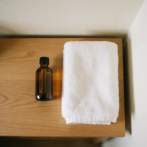 Bouteille en verre ambré contenant de l'huile de massage posée sur une serviette blanche pliée, le tout disposé sur une table en bois clair éclairée par une lumière naturelle douce.