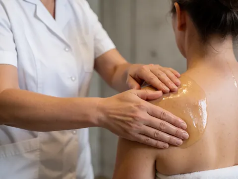 Mains expertes du thérapeute en mouvement appliquant de l'huile sur l'épaule du client. Le praticien porte un uniforme blanc dans un espace à la lumière chaude et au décor minimaliste.