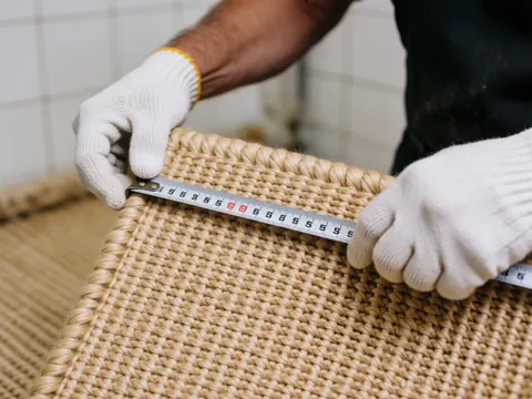 Une main tenant une règle métallique pour mesurer la largeur d'une moquette tissée de près, mettant en évidence la texture et la qualité du travail artisanal sur fond blanc.