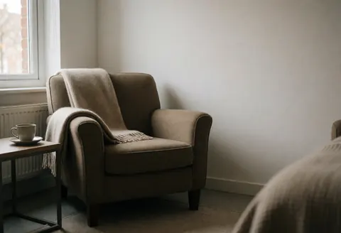 Un fauteuil confortable avec une couverture et une tasse posée sur une petite table près d'une fenêtre, créant une ambiance relaxante et chaleureuse.