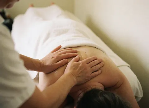 Gros plan des mains d'un professionnel massant l'épaule d'un client avec de l'huile, sur un fond blanc doux pour une ambiance relaxante.