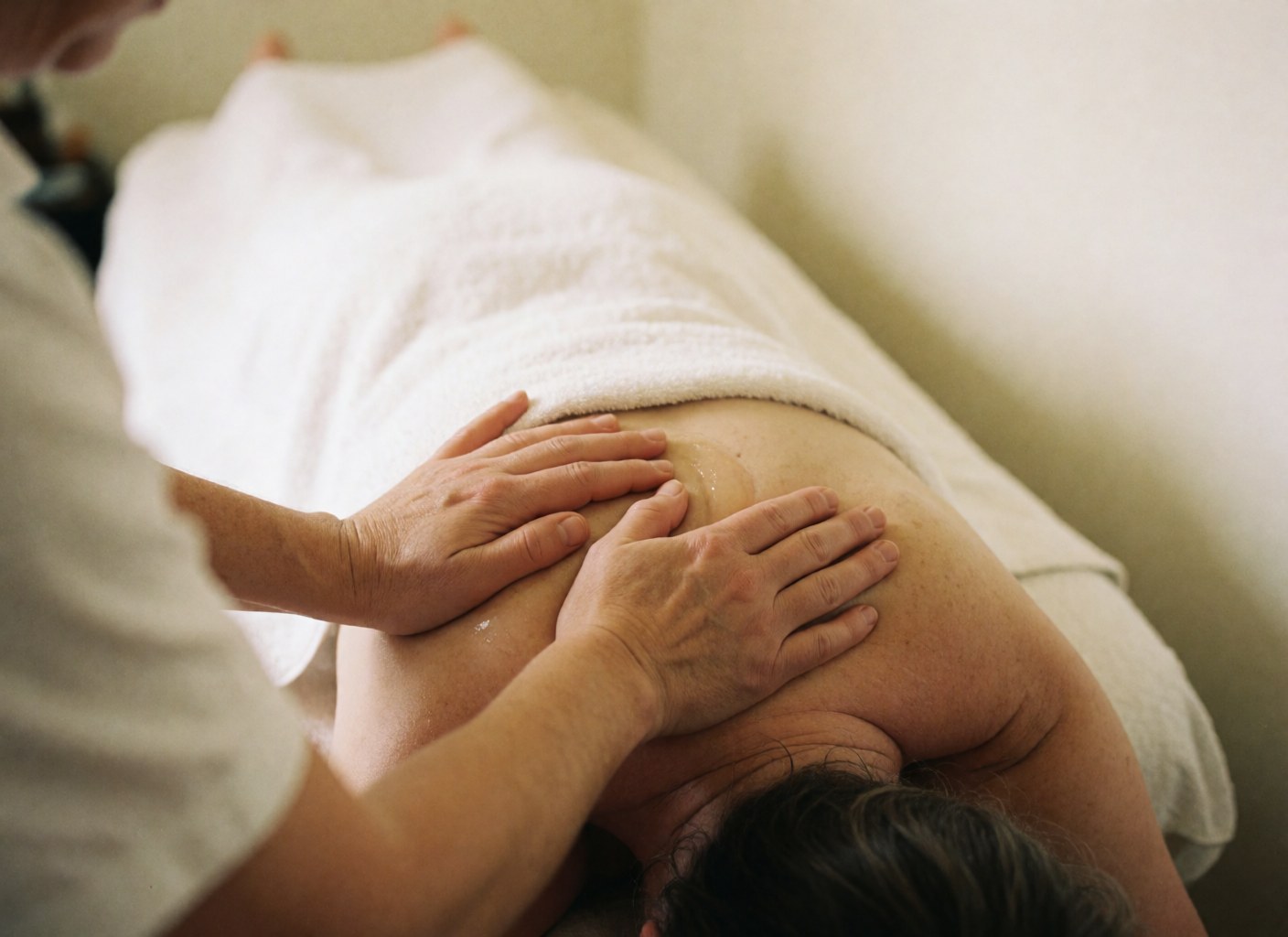 Gros plan des mains d'un professionnel massant l'épaule d'un client avec de l'huile, sur un fond blanc doux pour une ambiance relaxante.