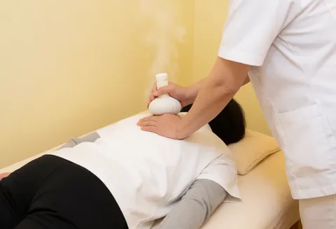 Un thérapeute en uniforme blanc utilise une boule d'herbes chaudes et vapeur sur le dos d'un client allongé sur une table de massage dans un cadre relaxant.