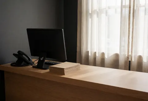 Bureau de réception avec ordinateur, téléphone et brochures, lumière douce par la fenêtre, murs gris, ambiance propre et ordonnée.