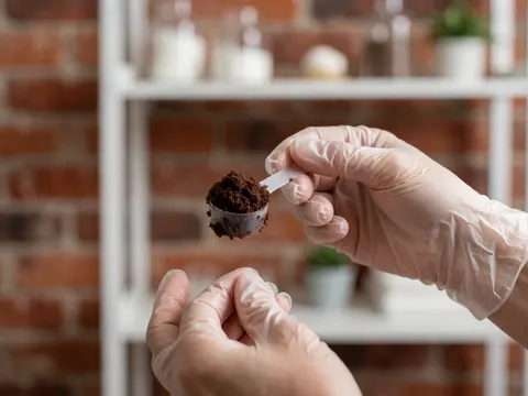Une main portant un gant tient une cuillère remplie d'exfoliant au chocolat devant un mur de briques et une étagère blanche.