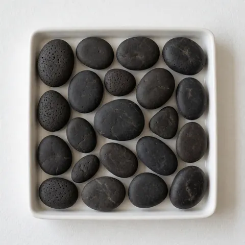 Roches volcaniques noires lisses et texturées disposées sur un plateau blanc, idéales pour les massages thaïlandais, les thérapies de relaxation et le bien-être naturel.