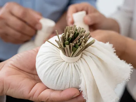 Gros plan d'une compresse herbale de massage thaïlandais avec des herbes sèches comme le citronnelle et les feuilles de kaffir, mains travaillant sur un tissu blanc, ambiance thérapeutique.