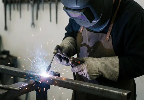 Un soudeur équipé d'un casque et de gants réalise une soudure sur une barre d'acier, des étincelles volent, des outils sont suspendus au mur blanc de l'atelier.