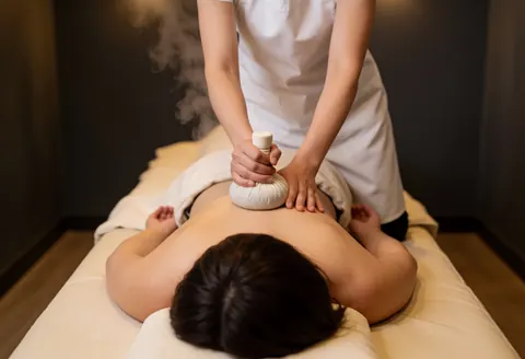 Un thérapeute applique un massage thaïlandais avec des compresses d'herbes sur le dos d'un client allongé, dans une ambiance chaleureuse et relaxante.
