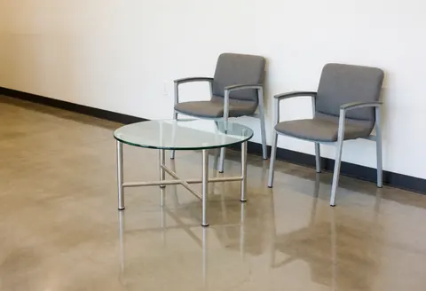 Coin d'attente contemporain doté de deux sièges gris et d'une table basse en verre posée sur un sol en béton poli, sous un éclairage fluorescent.