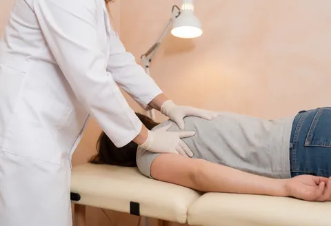 Un thérapeute professionnel en gants réalise une thérapie manuelle sur l'épaule d'un patient dans un cabinet aux murs de terre cuite et à la lumière chaude.