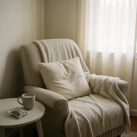 Un fauteuil d'appoint avec une couverture et un coussin, une tasse de thé sur une petite table près d'une fenêtre laissant entrer une lumière douce pour un environnement relaxant.